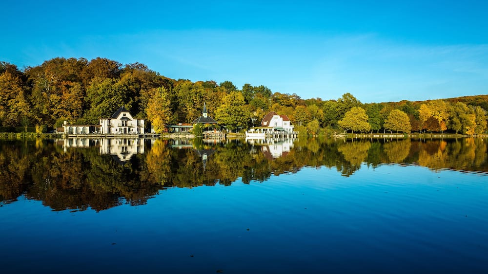 Un lac paisible où se reflètent les maisons et les arbres, le tout entouré d’un feuillage automnal.