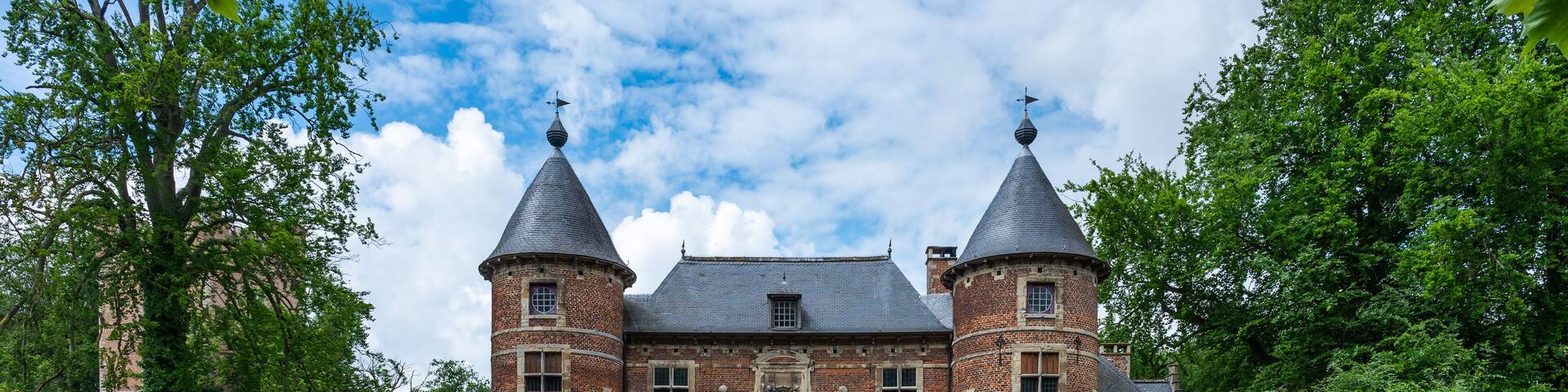 A historic brick castle with two towers, a cobblestone bridge, and lush greenery.