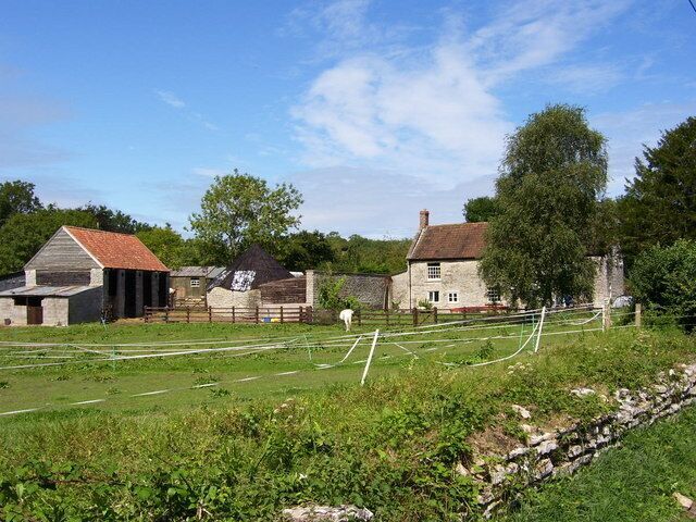 A rural landscape with a stone wall, a fenced field, and several old buildings.