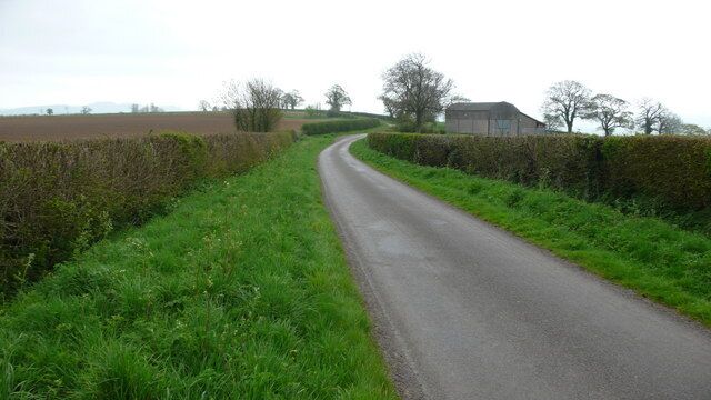 A narrow rural road curving through fields with hedgerows and a distant building.