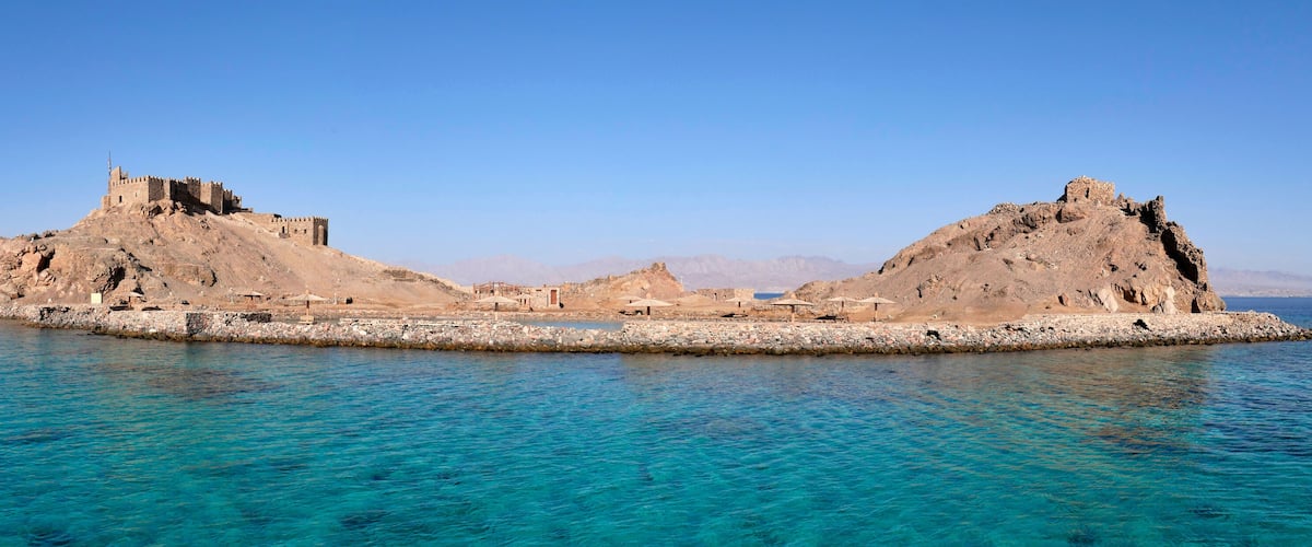 A coastal fortress with a prominent tower and a smaller building, surrounded by clear blue waters and rocky terrain.