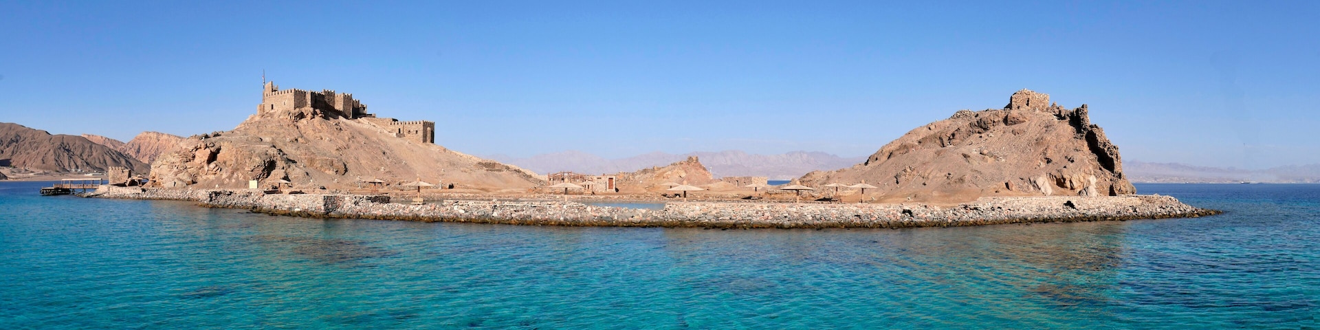 A coastal fortress with a prominent tower and a smaller building, surrounded by clear blue waters and rocky terrain.