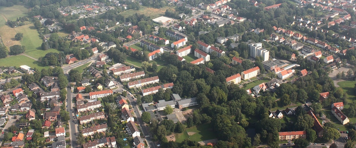 An aerial view of a densely populated urban area with numerous red-roofed residential buildings, green spaces, and a mix of commercial and industrial structures.