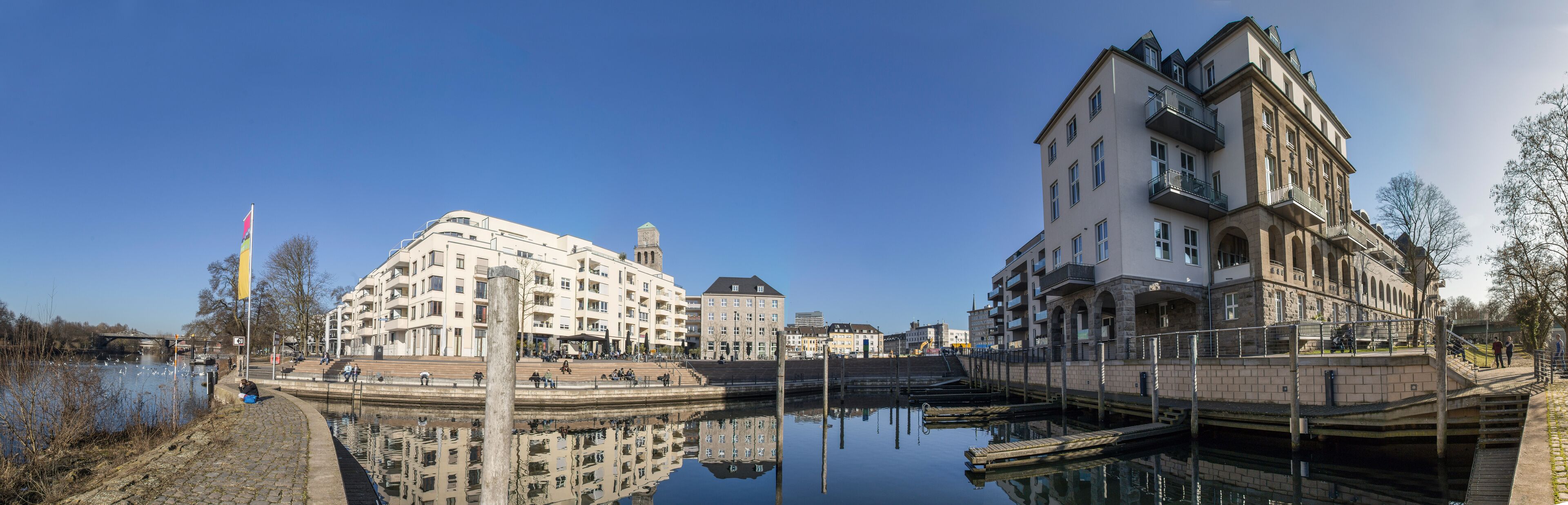 A riverside promenade with historic buildings, a bridge, and a calm water body reflecting the architecture.