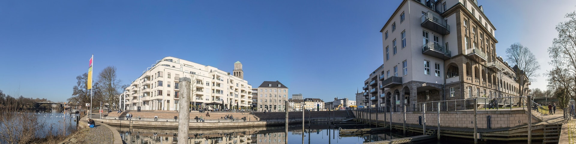 A riverside promenade with historic buildings, a bridge, and a calm water body reflecting the architecture.
