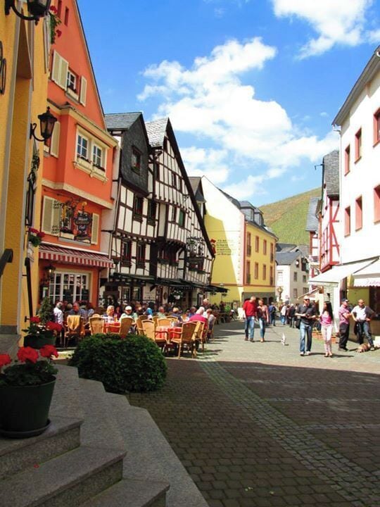 A cobblestone street with traditional half-timbered buildings, outdoor seating, and people walking.