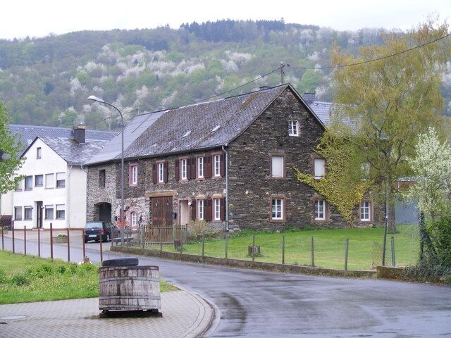 A stone building with a sloped roof, a paved road, and a grassy area with trees.