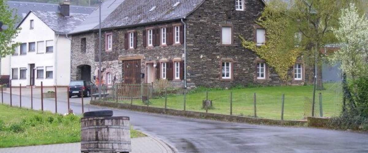 A stone building with a sloped roof, a paved road, and a grassy area with trees.