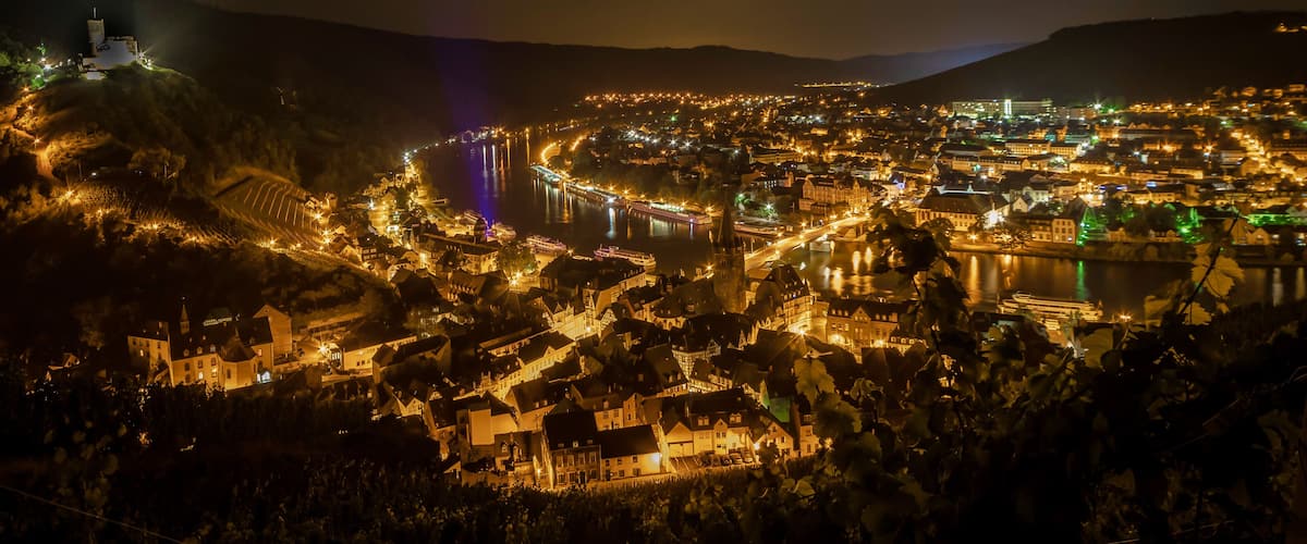 A nighttime view of a riverside town with illuminated buildings, a prominent church, and a bridge.