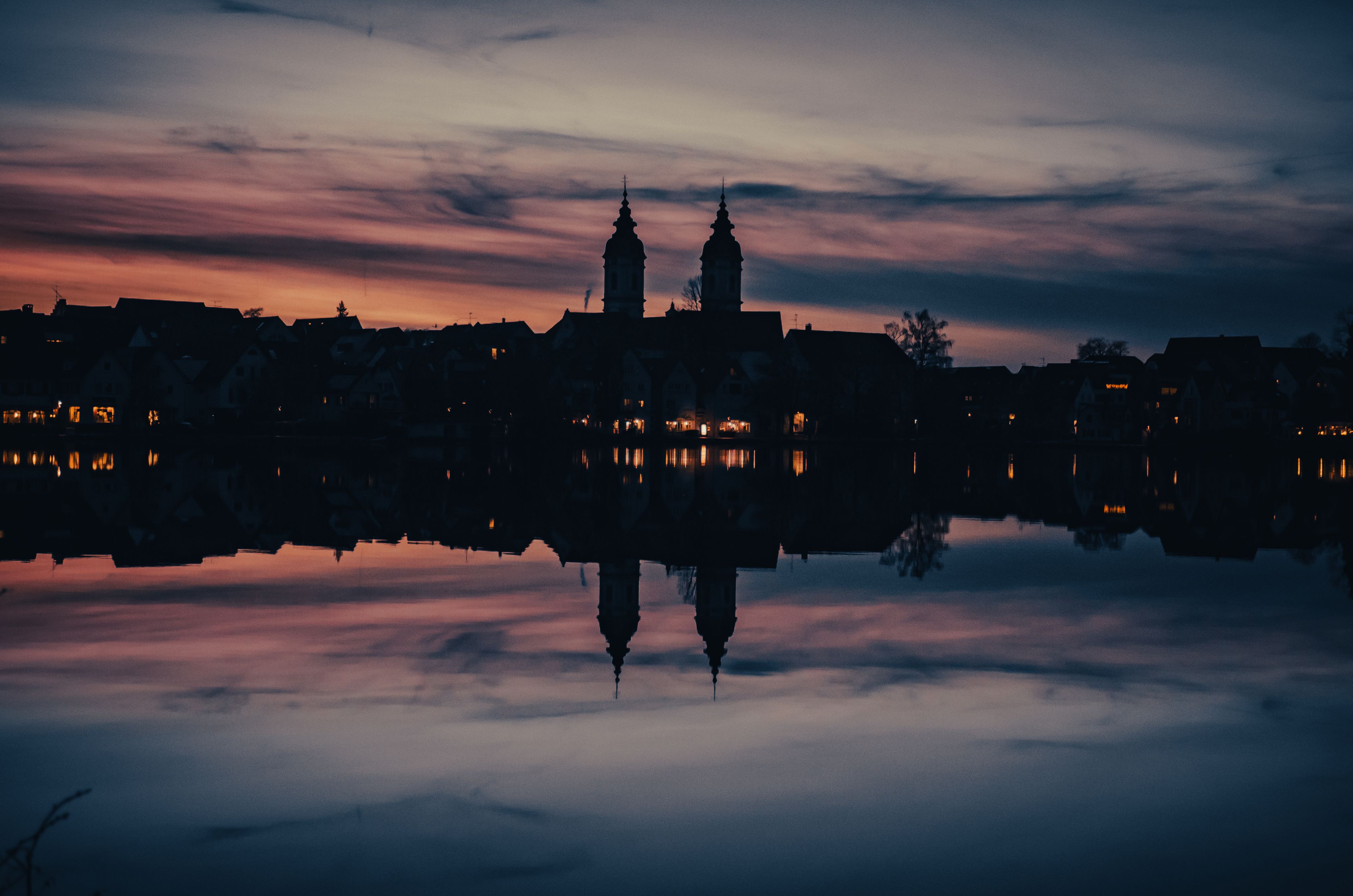 A city skyline at dusk with two prominent towers reflected in a calm body of water.