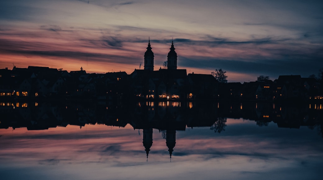 A city skyline at dusk with two prominent towers reflected in a calm body of water.