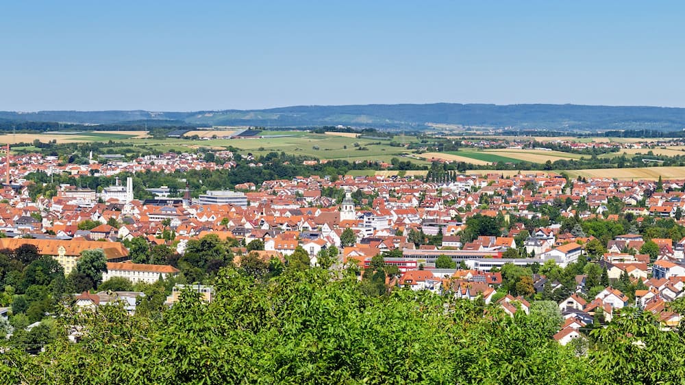 A town with red-roofed houses, green fields, and distant hills.