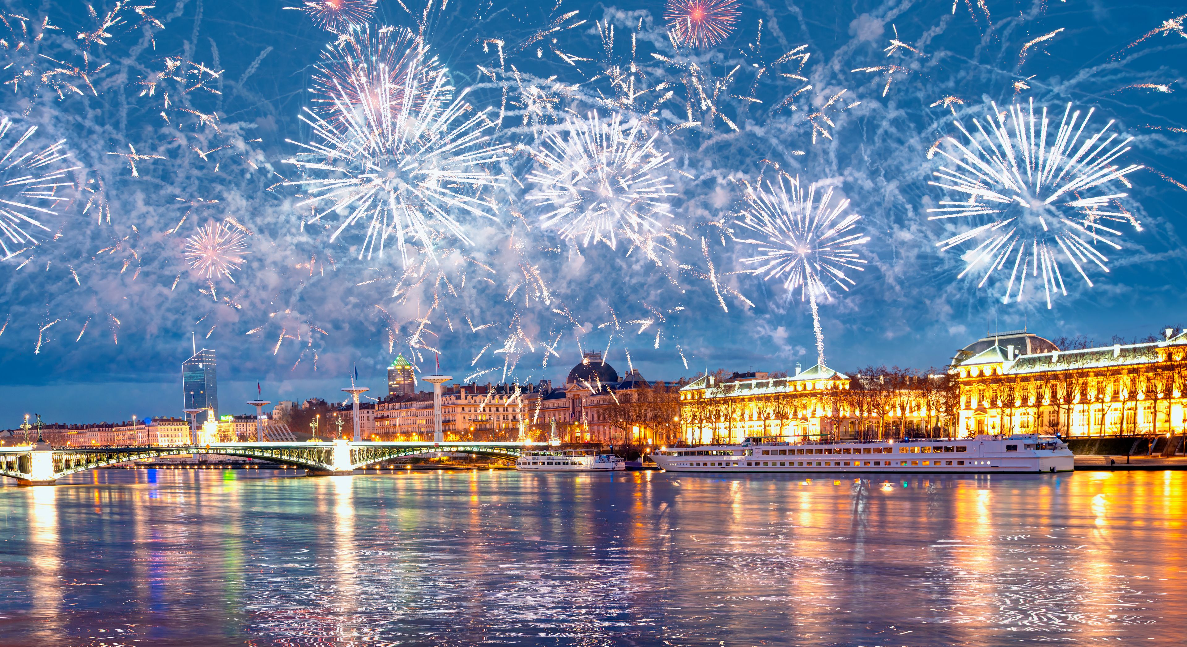Feux d’artifice au-dessus d’une rivière, avec un pont et des bâtiments en arrière-plan.