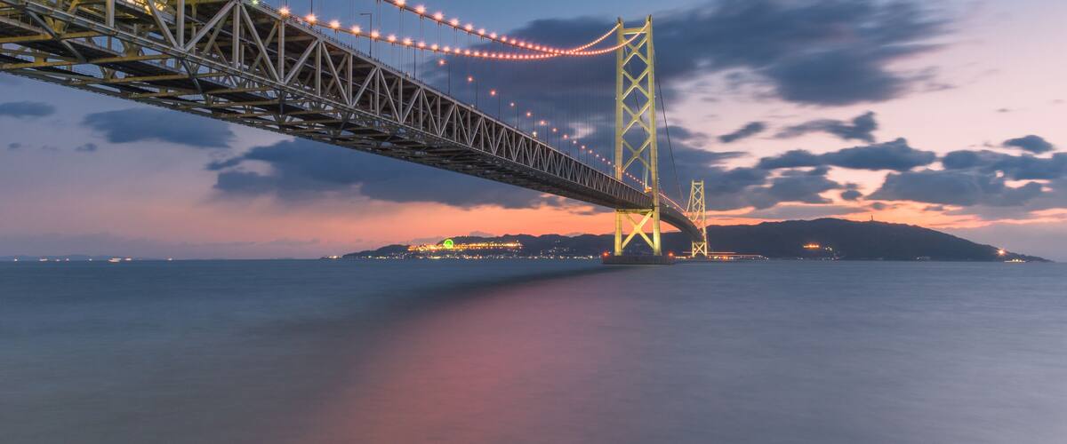 A bridge with lights, a calm sea, and a sunset sky.