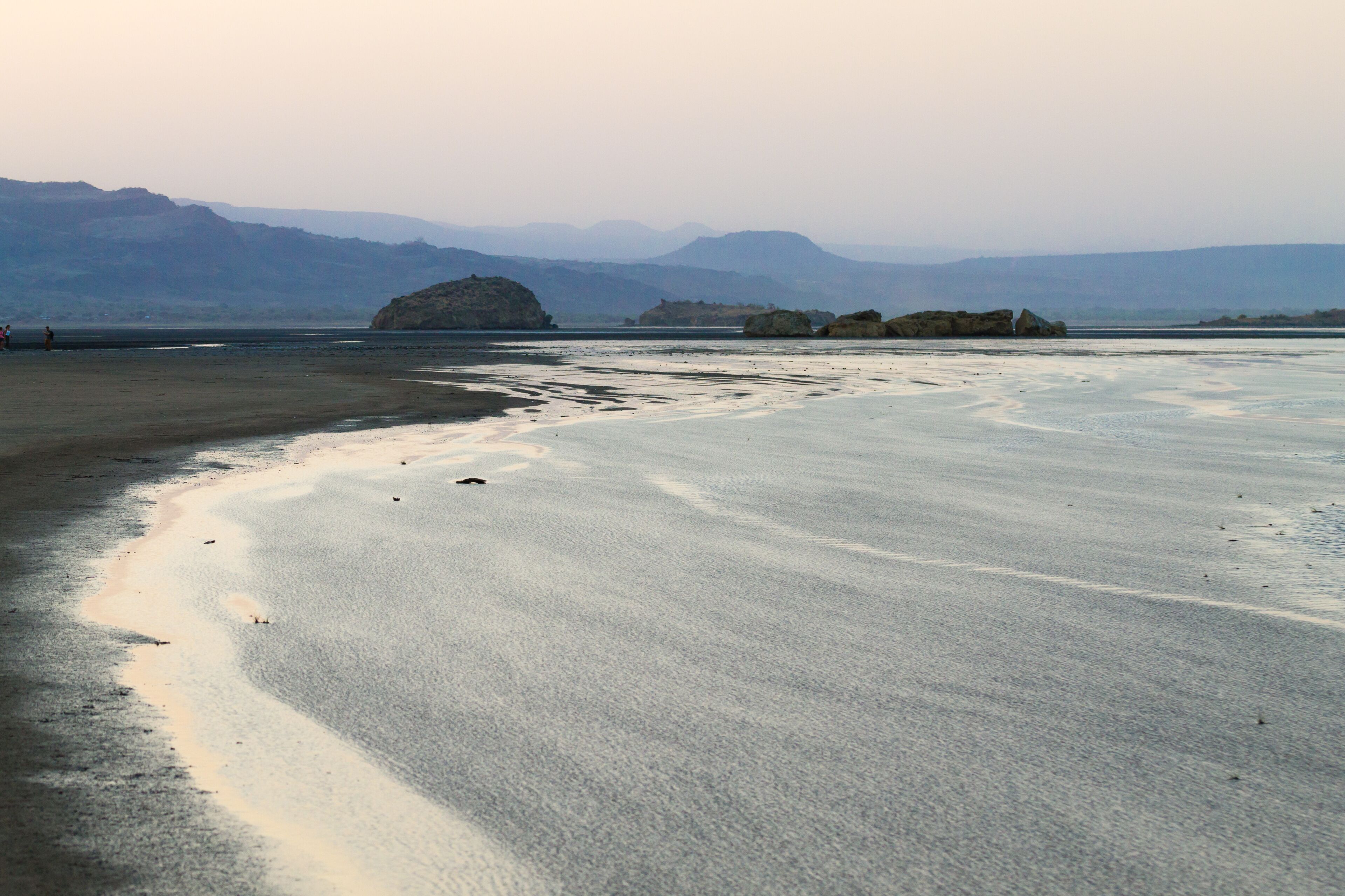 A beach with calm waters, distant mountains, and a clear sky.