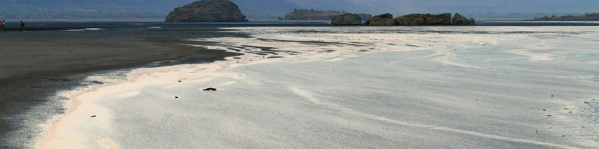 Una playa con aguas tranquilas, montañas a lo lejos y cielo despejado.