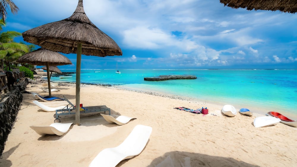 Ein Strand mit StrohdĂ€chern, LiegestĂŒhlen und einem ruhigen tĂŒrkisfarbenen Meer.