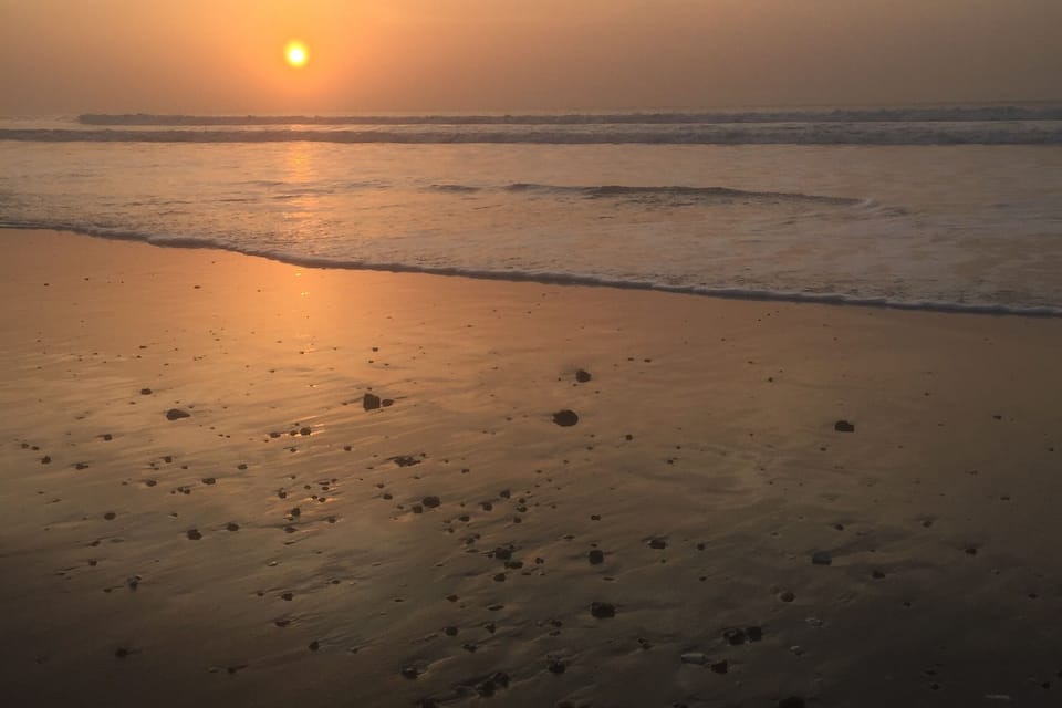 Een strand bij zonsondergang, de zon laag aan de horizon, golven die zachtjes op het strand komen en voetsporen in het zand.