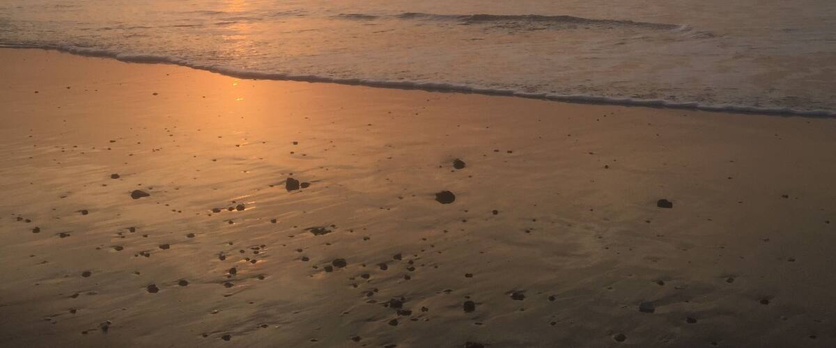 A beach at sunset with the sun low on the horizon, waves gently lapping the shore, and footprints in the sand.