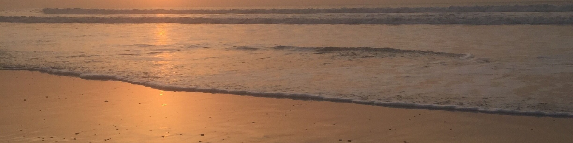 A beach at sunset with the sun low on the horizon, waves gently lapping the shore, and footprints in the sand.