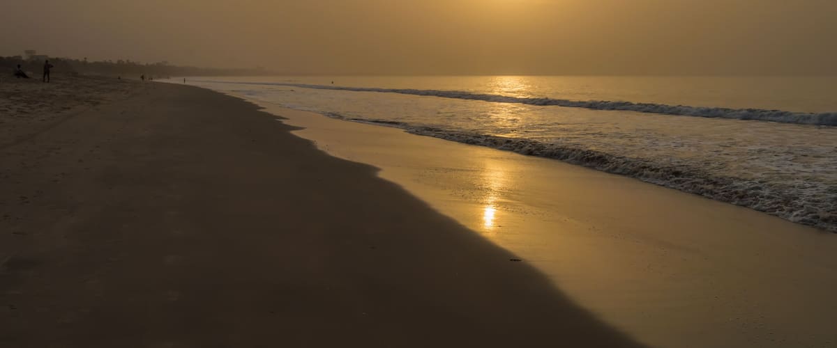 A beach with a calm sea, a setting sun, and a few people walking along the shore.