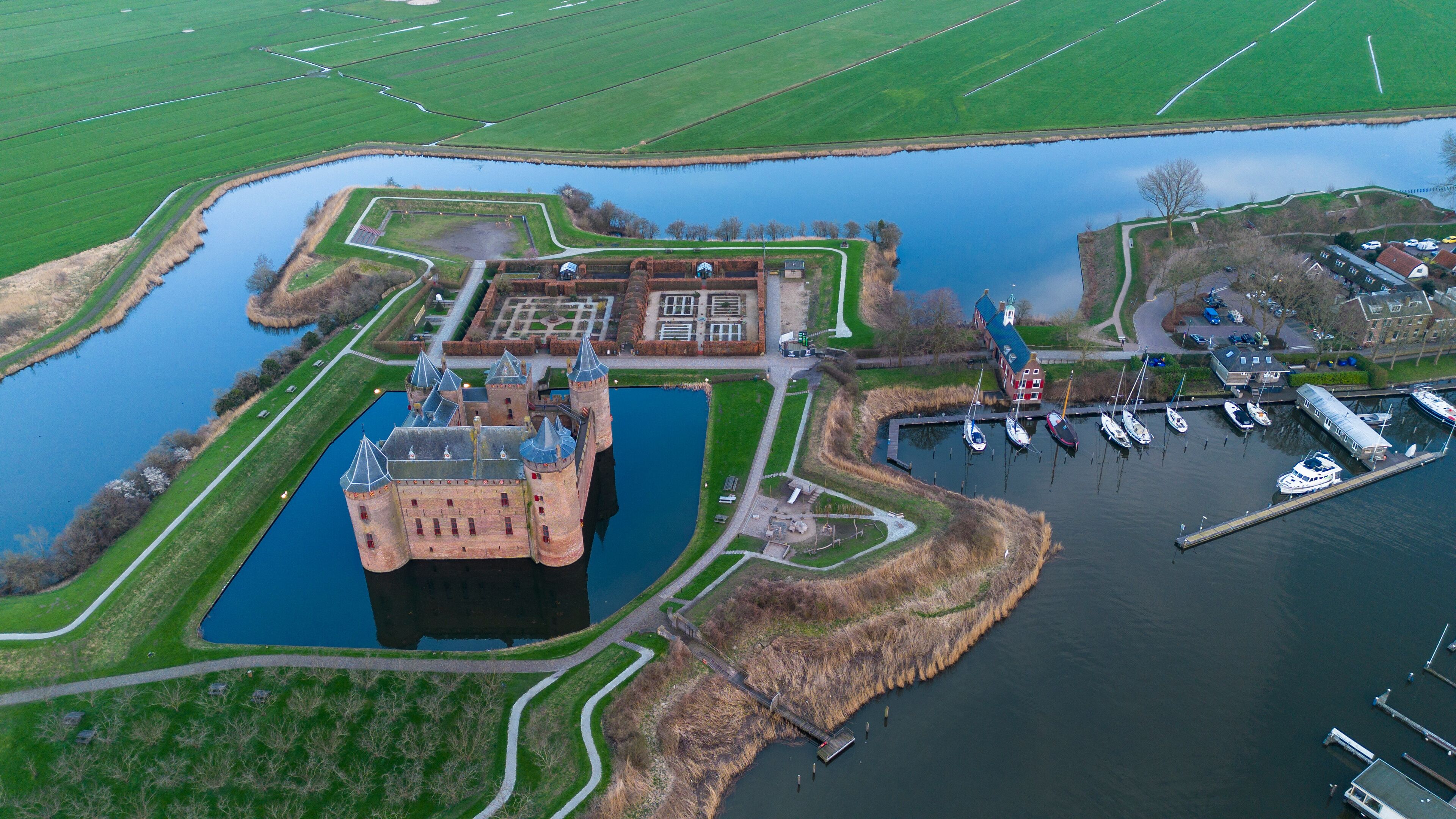 A castle surrounded by water, a marina with boats, and green fields.