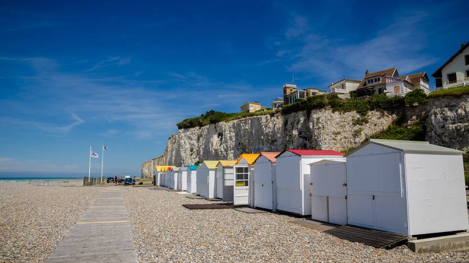 A pebble beach with colorful beach huts, a wooden boardwalk, and houses on a cliff.