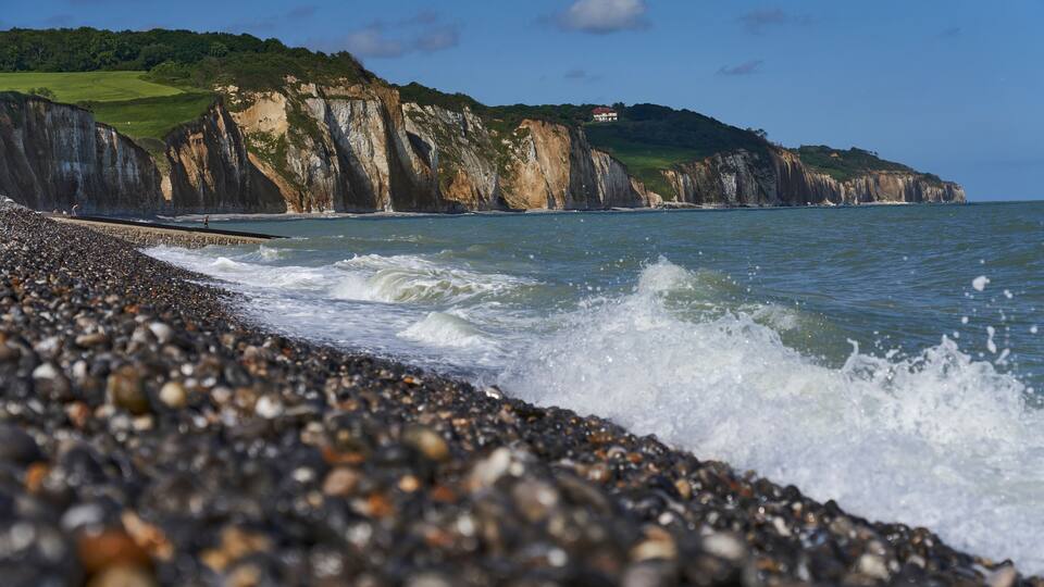 A pebbled beach with waves crashing onto the shore, cliffs in the background, and a house on a hill.