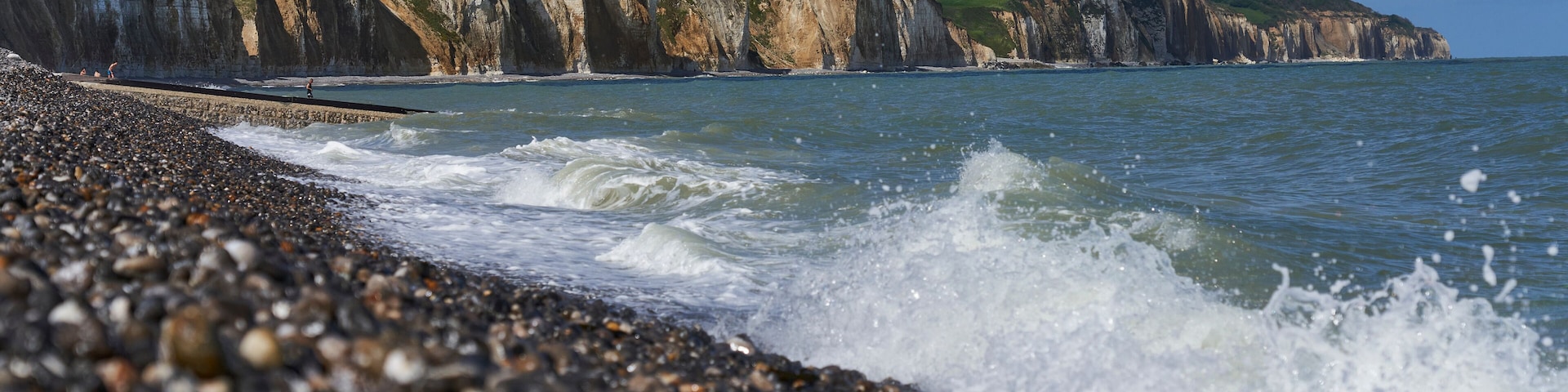 Una playa de guijarros con olas rompiendo en la orilla, acantilados al fondo y una casa en una colina.