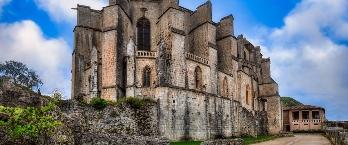 A historic stone castle with multiple towers and arched windows, surrounded by greenery and a clear sky.