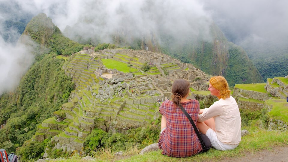 Two people sitting on a grassy ledge overlooking Machu Picchu ruins.
