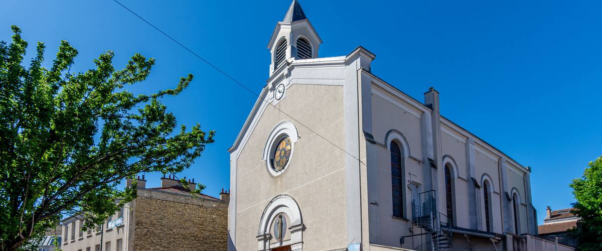 A church with a clock tower, a stone building, and a street with cars and trees.