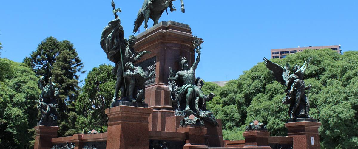 A bronze equestrian statue with multiple figures, set against a backdrop of trees and a clear blue sky.