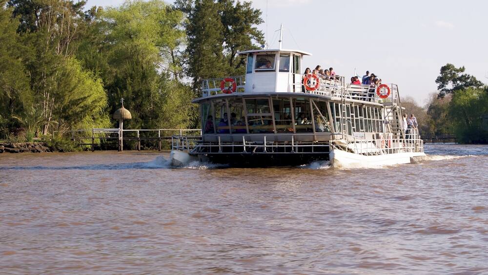 A riverboat with passengers, surrounded by trees and a calm water surface.