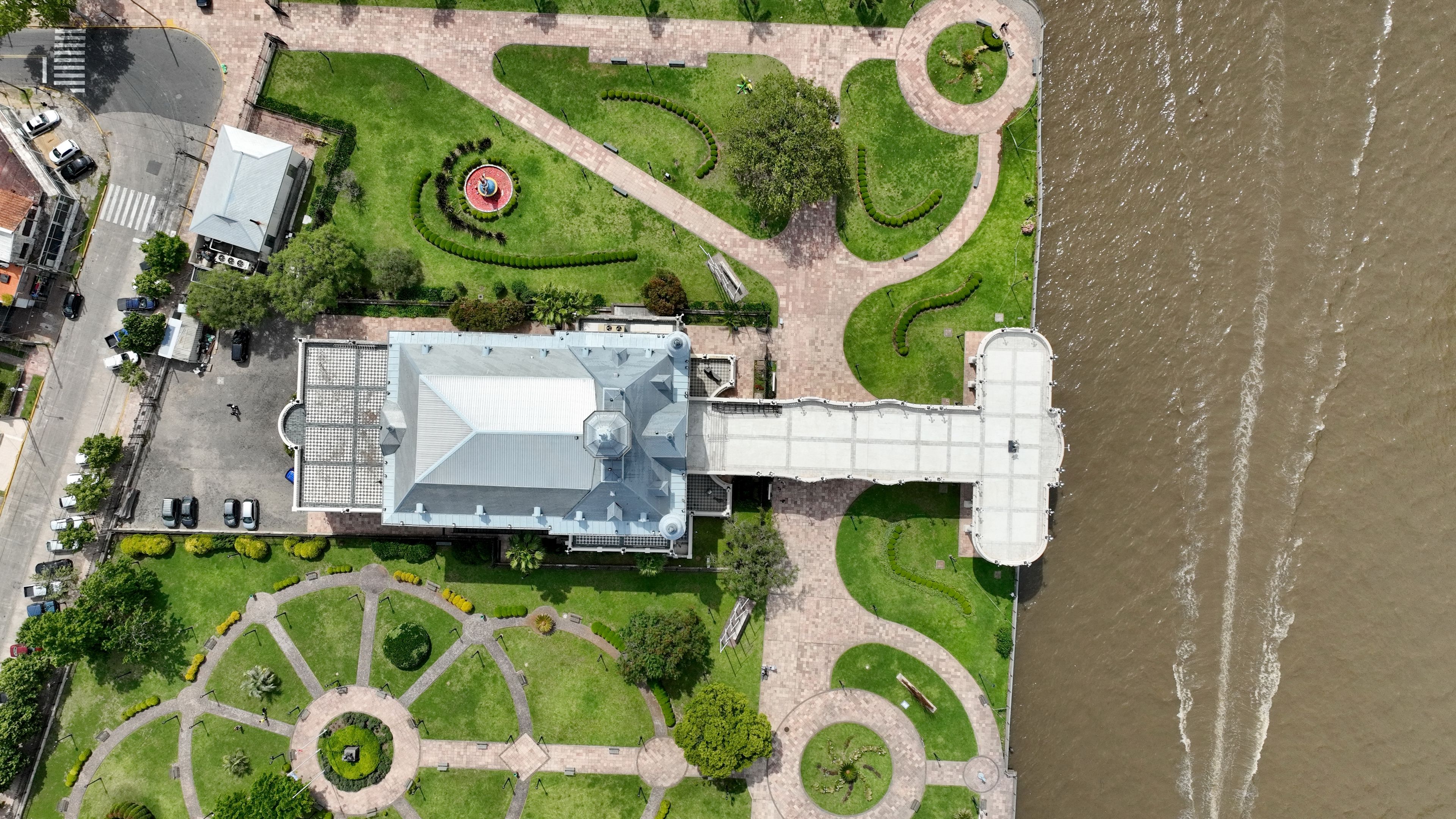 Luftaufnahme eines Flussuferparks mit einem großen Gebäude, Wegen und Grünflächen.