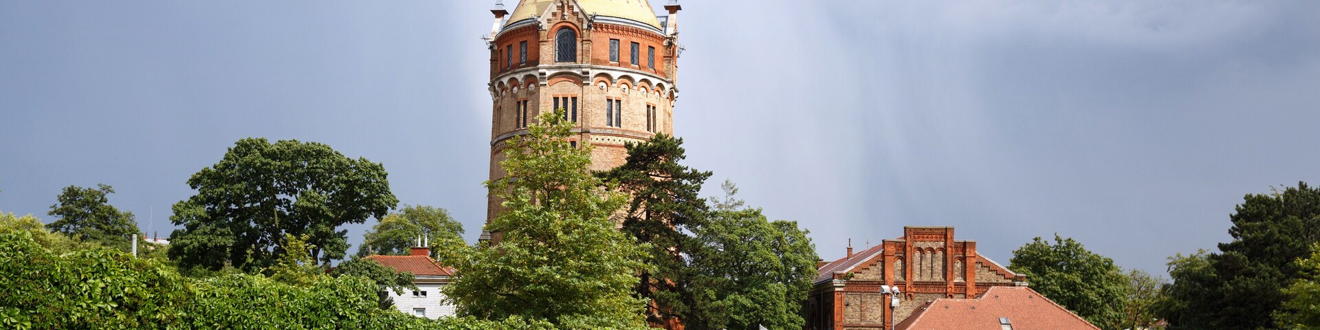 Uma torre histórica com cúpula e pináculo, rodeada por um jardim bem cuidado com arcos cobertos de hera.