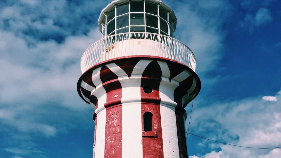 A red and white lighthouse with a weather vane on top, set against a partly cloudy sky.
