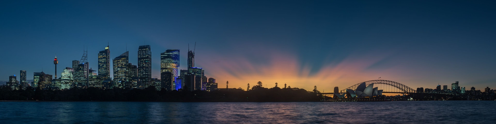 Une vue sur les gratte-ciel d’une ville au crépuscule, avec des bâtiments illuminés et l’Opéra de Sydney en arrière-plan.