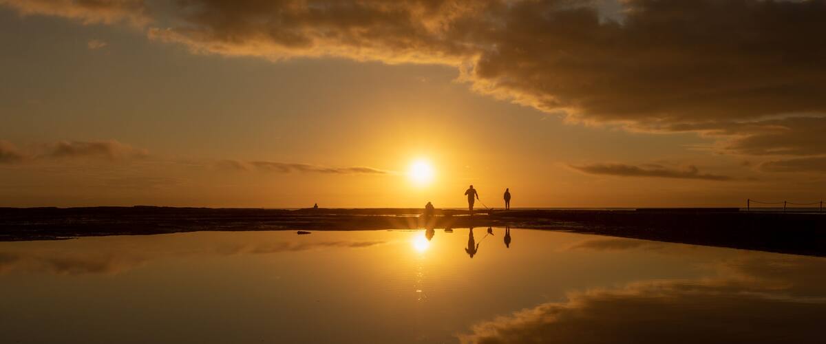 A sunset over a reflective water surface with silhouettes of people and a distant shoreline.