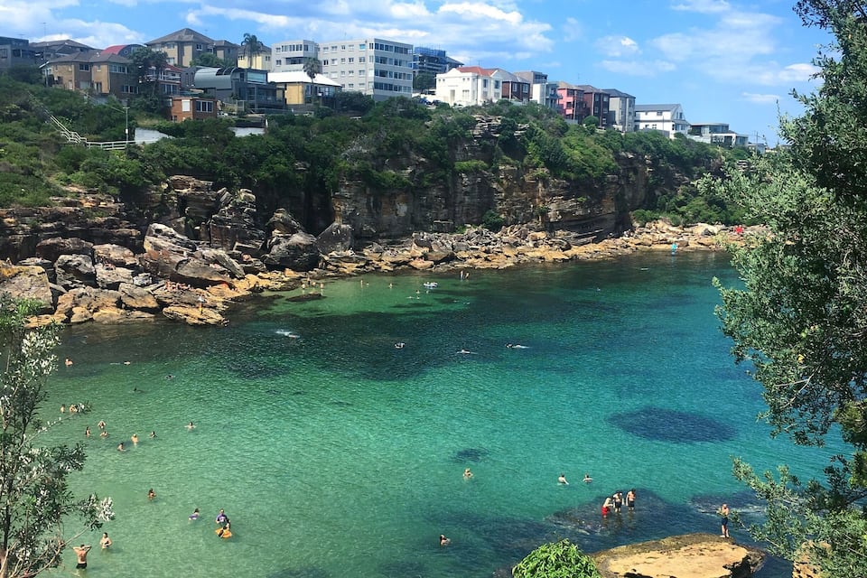 A coastal area with clear turquoise water, rocky shorelines, and buildings on a hillside.