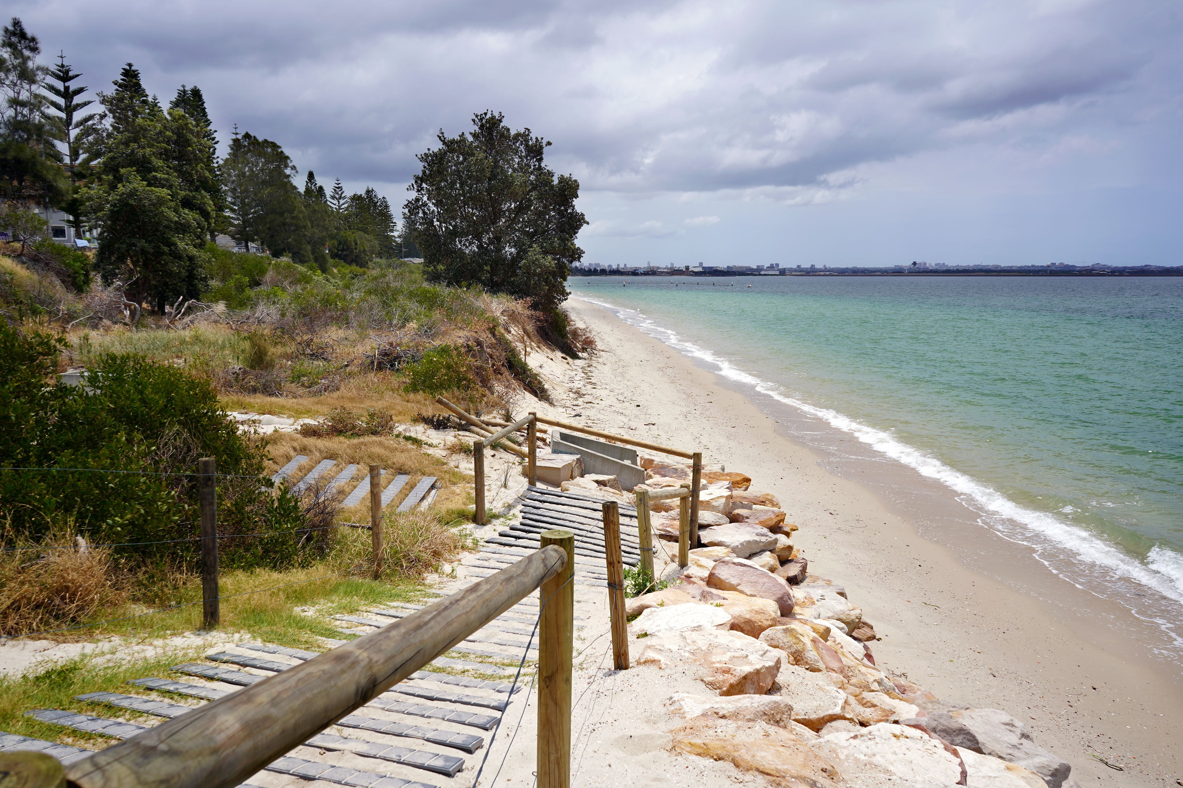 A wooden staircase leading down to a sandy beach with a rocky shoreline, surrounded by trees and a cloudy sky.