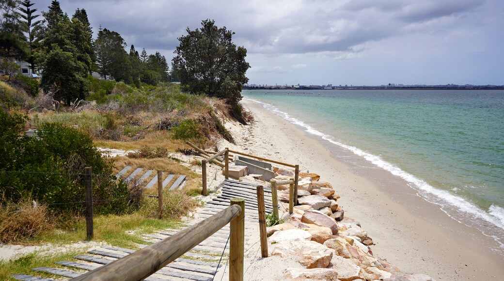 A wooden staircase leading down to a sandy beach with a rocky shoreline, surrounded by trees and a cloudy sky.