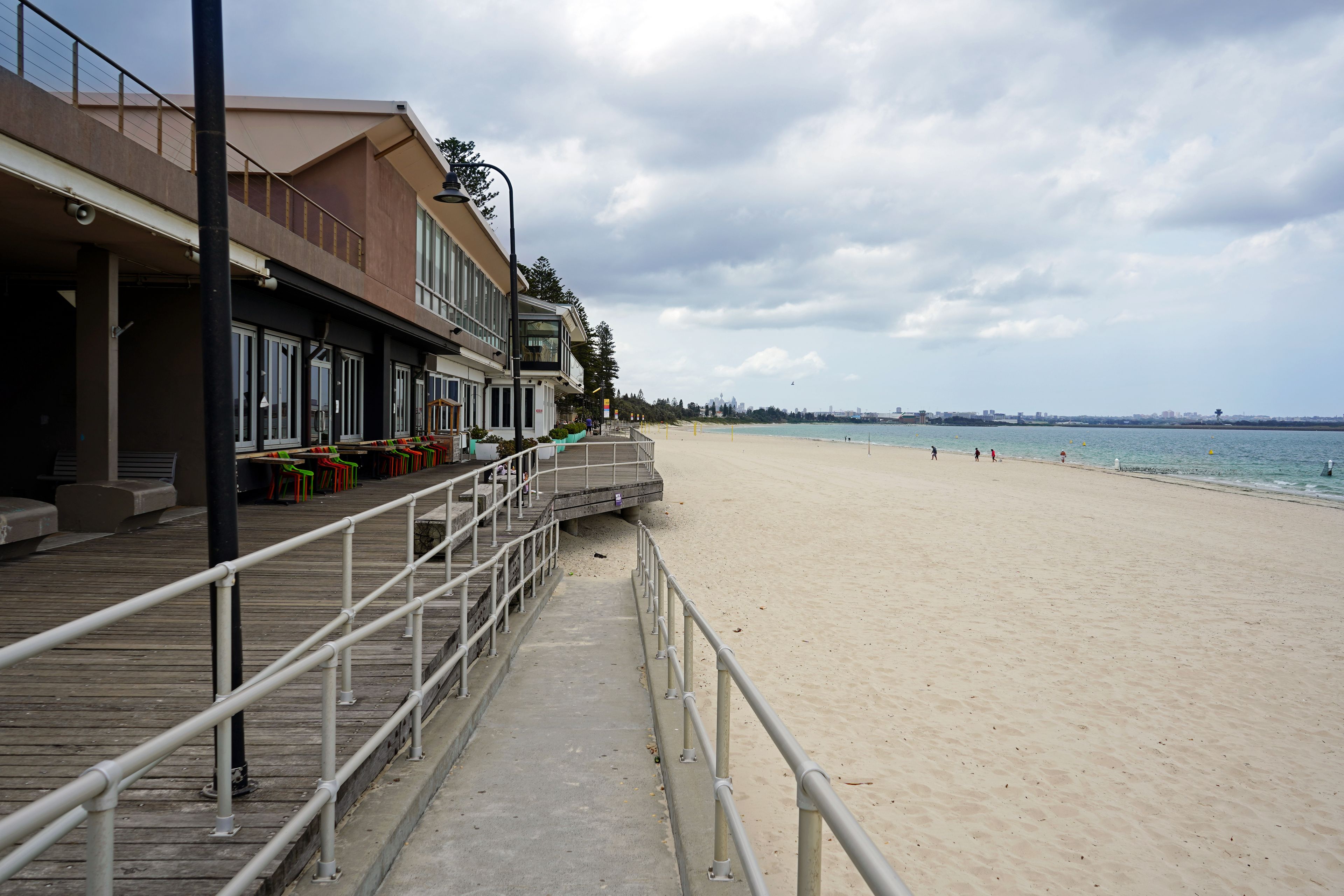 A beach with a wooden boardwalk, a building with large windows, and a city skyline in the distance.