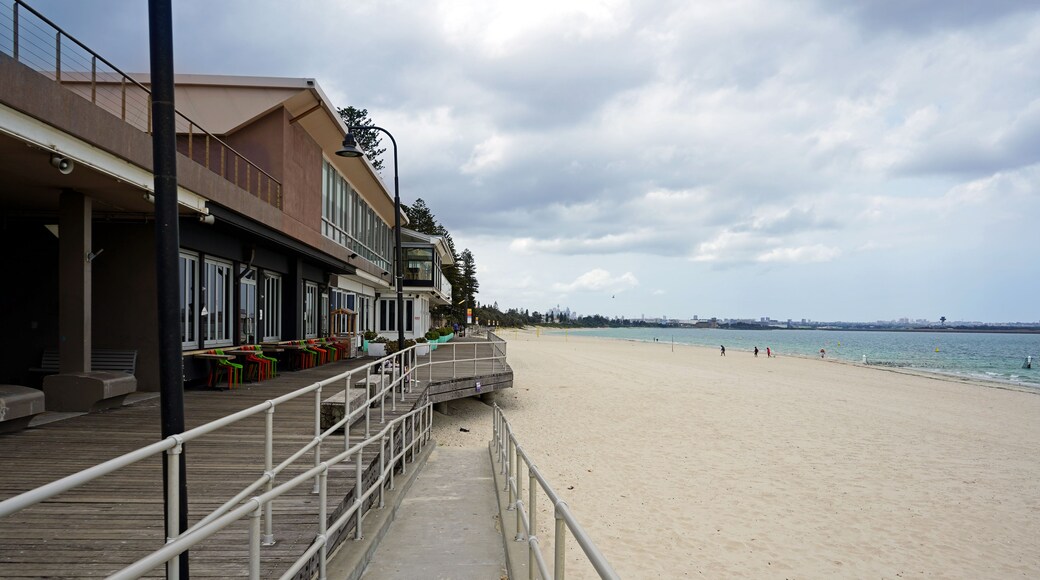 A beach with a wooden boardwalk, a building with large windows, and a city skyline in the distance.