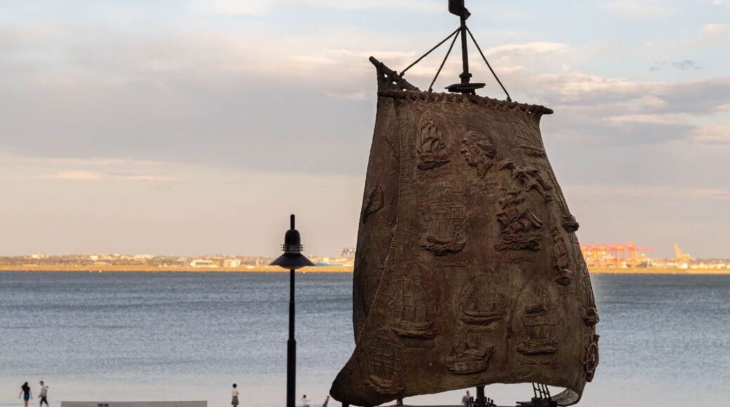A large, sail-shaped sculpture with ship carvings and a figurehead, mounted on a pole, by a waterfront with a distant cityscape.