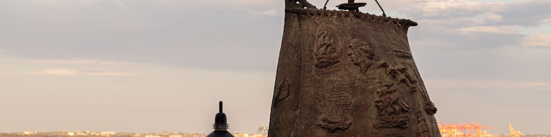 A large, sail-shaped sculpture with ship carvings and a figurehead, mounted on a pole, by a waterfront with a distant cityscape.