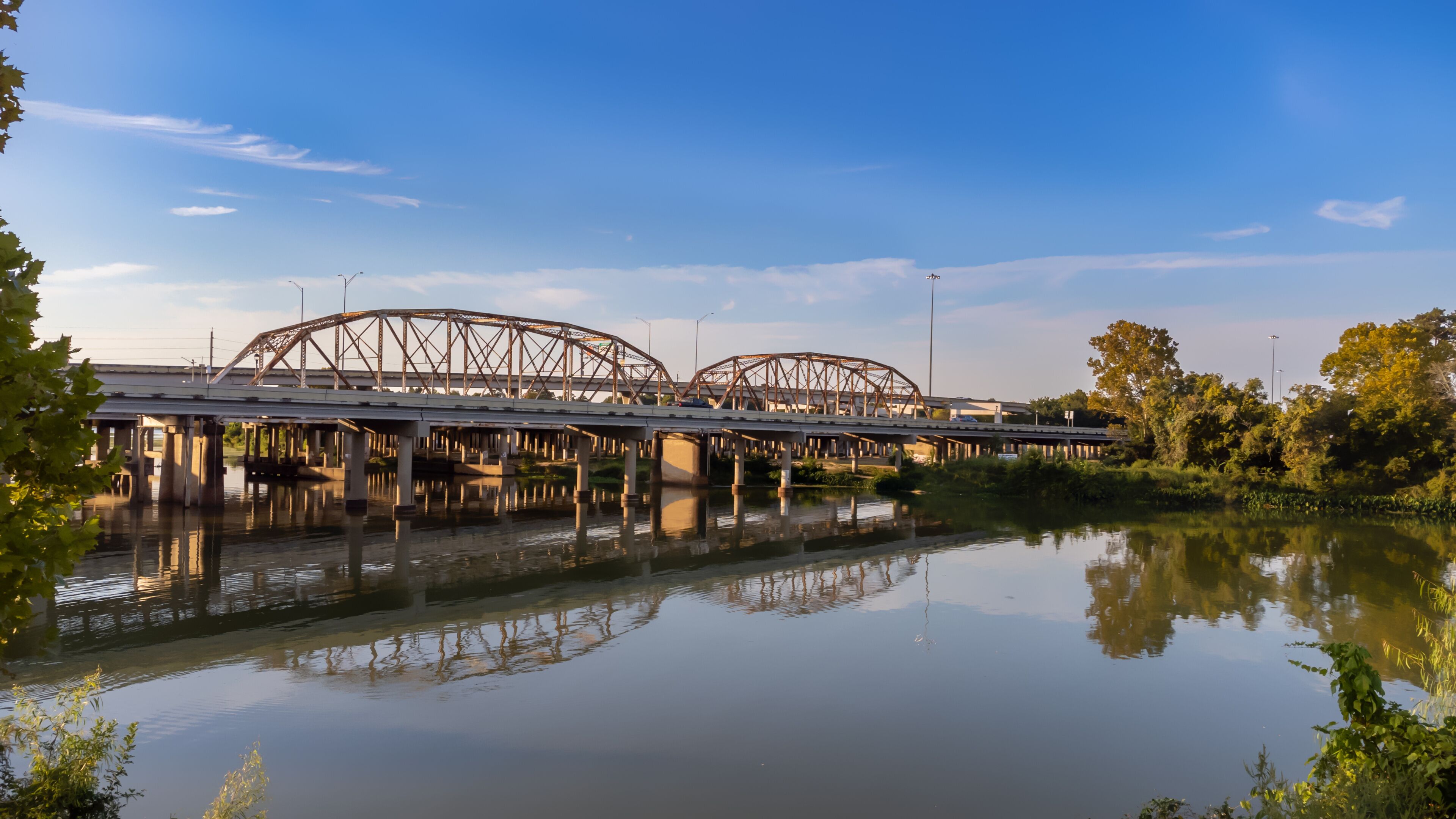 A steel bridge with arches spanning over a calm river, surrounded by greenery.