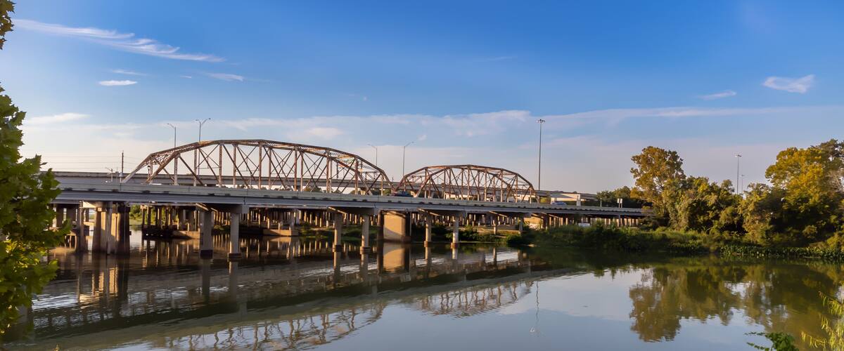A steel bridge with arches spanning over a calm river, surrounded by greenery.