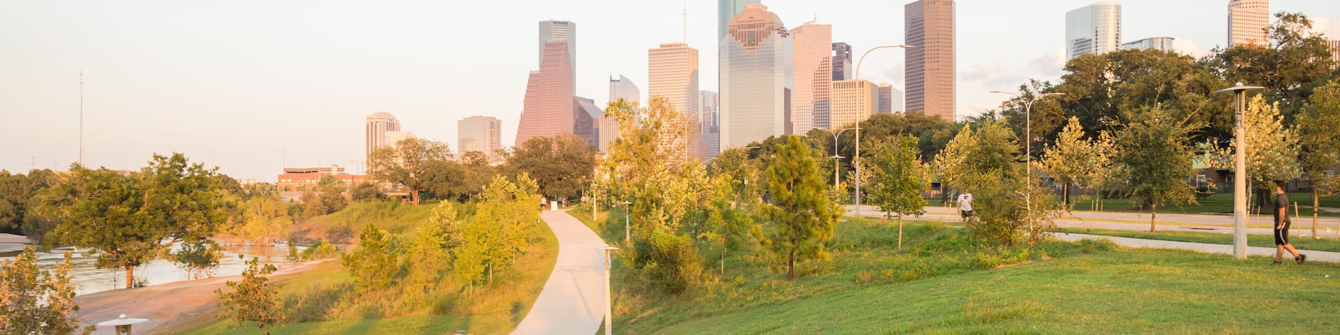 Panorama of downtown Houston, Texas, USA at sunset from Eleanor Tinsley Park. Grassy green park lawn, Buffalo Bayou river, curved pathway with people walking, biking, exercising and modern skylines.