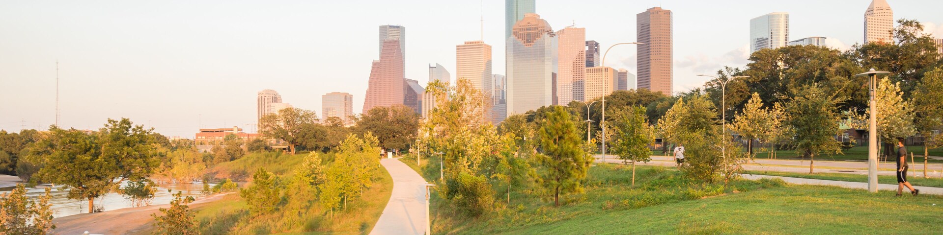 Panorama of downtown Houston, Texas, USA at sunset from Eleanor Tinsley Park. Grassy green park lawn, Buffalo Bayou river, curved pathway with people walking, biking, exercising and modern skylines.